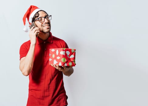 Smiling guy in christmas hat holding christmas gift box and talking on the phone. Christmas man holding gift and calling family on phone. Happy man holding gift and calling family at christmas