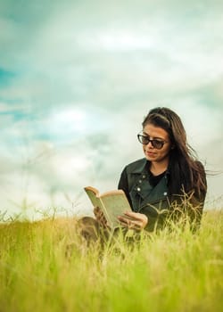 Smiling girl reading a book in the green field, Teen girl sitting on the grass reading a book. Concept of girl in glasses reading in the field, Attractive young woman reading a book in the field
