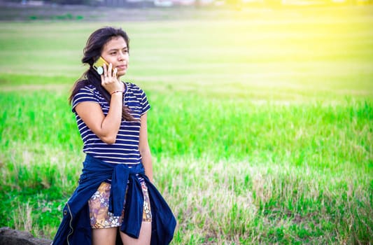 Beautiful young woman calling by cell phone on the grass outdoor. Young girl calling phone on the green field