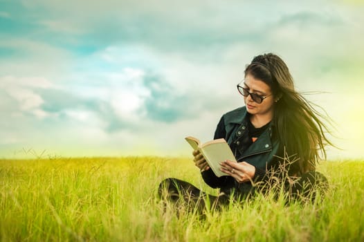Attractive young woman reading a book in the field, Smiling girl reading a book in the green field, Teen girl sitting on the grass reading a book. Concept of girl in glasses reading in the field