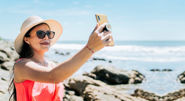 Girl in hat on the beach taking a selfie. Young woman on vacation taking photos on the beach, Smiling girl in hat taking photos on the beach. Beach vacation concept
