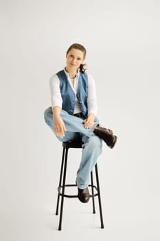 Stylish woman in denim vest and wide jeans sitting on a high chair over grey background