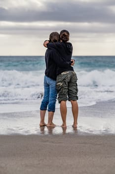 Mother and daughter standing barefoot on the foreshore intensely embracing each other while observing the horizon over the sea. A young mother spending leisure time with her teenage daughter, conceptual theme concerning family relationships.