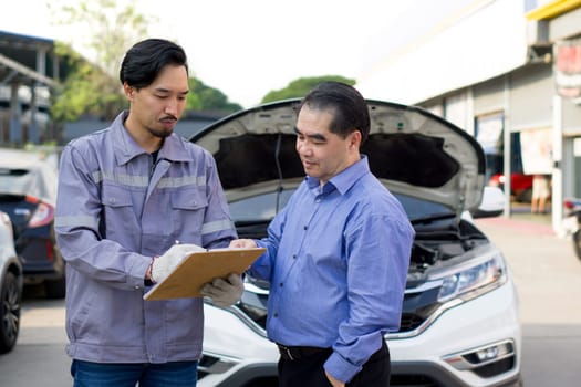 A mechanic in work uniform with gloves, holding document clipboard focused on the paperwork. The car owner or a service advisor discussing the vehicle's issues or maintenance work with the technician