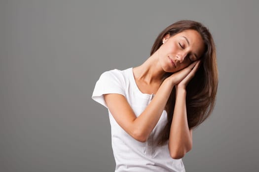 Beautiful young woman with long hair mimics sleeping gesture, hands joined, pretending to sleep on hand-formed pillow, tilting head to one side, conveying calmness and serenity