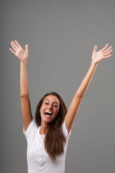 Frontal emotional portrait of a young woman isolated on a neutral background. She's overjoyed, shouting YEEE with open mouth, raising arms and open hands with 10 fingers. Long brown hair, slim, white