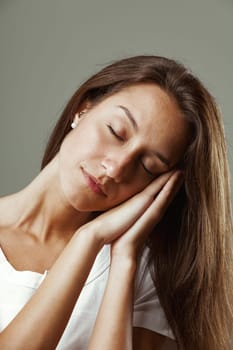 Beautiful young woman with long hair mimics sleeping gesture, hands joined, pretending to sleep on hand-formed pillow, tilting head to one side, conveying calmness and serenity