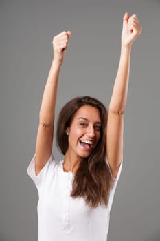 Emotional portrait of a young woman isolated on a neutral background. Arms raised straight up, hands clenched in fists, expression of joy and exultation. Open mouth and perfect teeth. Long brown hair