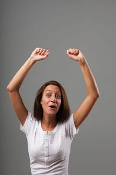 Emotional portrait of a young woman isolated on a neutral background. Exultant because she won something or succeeded. Hands raised in fists, mouth forms an 'OH.' Long brown hair, slim, white shirt. S