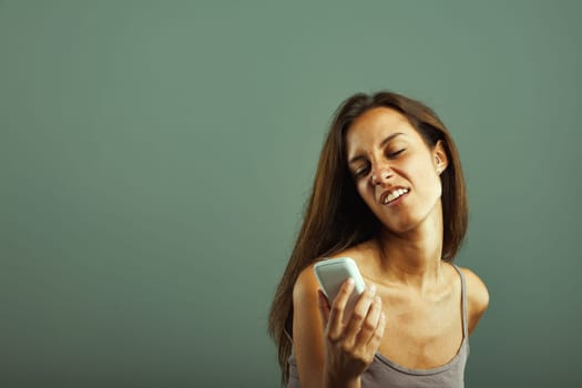 Emotional portrait of young woman, annoyed, mocking phone. Mimics person she dislikes. Envious, impulsive. Brown hair, slim, beautiful, beige tank top. State of mind concept