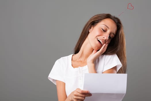 Emotional portrait of a young woman isolated on a neutral background. She tilts her head and places her hand on her cheek, touched and tender by the love expressed in the letter she's reading, overwhe