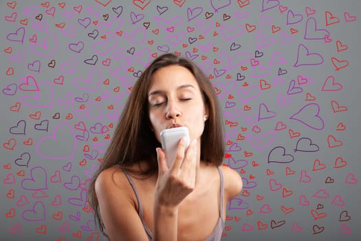 Young woman sends kisses while closing her eyes to her smartphone surrounded by hundreds of colorful hearts - concept of cell phone and love for Valentine's Day