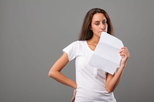 Emotional portrait of a young woman on a neutral background. Thoughtful girl, negative expression, disappointed and silently furious. Possibly sad about the letter she holds. Long brown hair, slim, wh