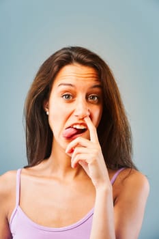 Emotional portrait of young woman isolated on neutral background. Girl puts finger in nose, makes a face. Immature response for laughs or because others don't deserve better. Long brown hair, slim.