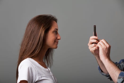 Long-haired girl in front of vintage compact camera lens held by older male hand. Slightly shy, unsure, awaiting instructions for a good photo. Side profile, white shirt