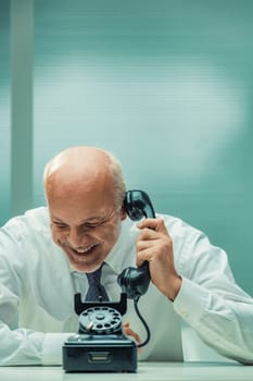 Overwhelmed by laughter, a mature businessman crouches with a phone, pointing to an antique rotary device. The conversation sparks immense amusement