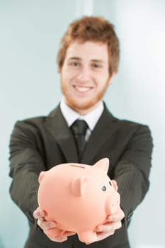 Close-up of piggy bank, held in hand and leaning forward by young businessman blurred in background. He smiles and considers that savings and investment are the basis of good business.