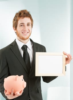 Copyspace by savings concept. Young smiling businessman holds a white sign available for your writing in one hand and a piggy bank symbolizing money and savings in the other.