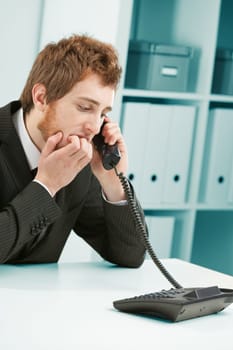 Fear, anxiety, communication stress? Worried-looking young employee bites his nails as he waits or listens at the handset of his office landline. either a mess has happened or he will be fired.