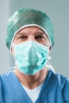Surgeon in close-up wears blue gown, mask, and cap. He looks serious, professional, around fifty, but smiles confidently