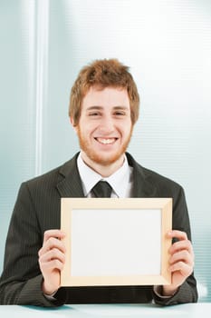 Young man very smiling with sign in hand sitting at a desk. He is in an office, business or IT workplace. The sign is a blank space for your COPYSPACE and writing.