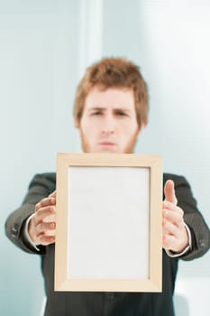 COPYSPACE in a blank, wood-framed sign in the foreground, held by two hands of a young employee. The man is in the background, blurred, and still has a negative, sad, blaming expression.