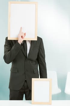 Sign-headed man framed in an office, he is standing with his finger in front of his face making the gesture of silence. On the desk in front of him is another smaller frame. COPYSPACE ready for surrea