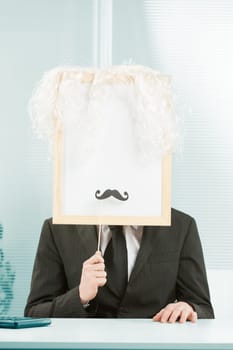 Man with framed sign head in an office. The sign-face also has a white wig and fake mustache added and is ready for you with COPYSPACE.