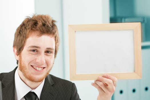 Young man very smiling with sign in hand sitting at a desk. He is in an office, business or IT workplace. The sign is a blank space for your COPYSPACE and writing.