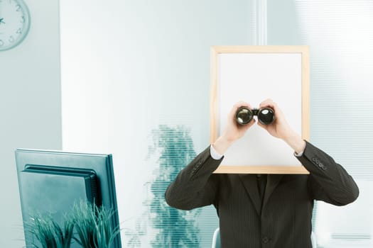 Man with sign head framed in an office where a computer monitor can also be seen. The man is standing up and watching you with binoculars. White space is free for editing.