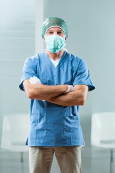 Proudly standing, a sturdy healthcare worker in hospital attire, blue top, white gloves, cap, and surgical mask. A fit middle-aged man, passionate about his job, smiles at you