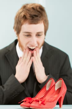 Man longs for those red shoes with white polka dots from women's heels. Young man (out of focus) with funny expression stares at shoes (in focus) on table with enthusiastic admiration. is dressed in o