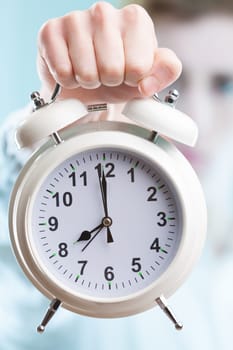 An in-focus hand extending a large alarm clock against a blurry office background. In business, time is money, and deadlines are just around the corner
