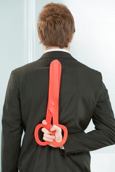 A clerk in black pinstripes in the office seen from behind. He hides a red scissor behind his back. It symbolizes that he is keeping future cost and personnel cuts or a general danger in a state of cr