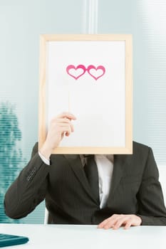 Man with framed sign head in an office. In front of the fake head made of signboard he also puts cardboard glasses in the shape of pink hearts to complete a disguise that YOU can customize graphically
