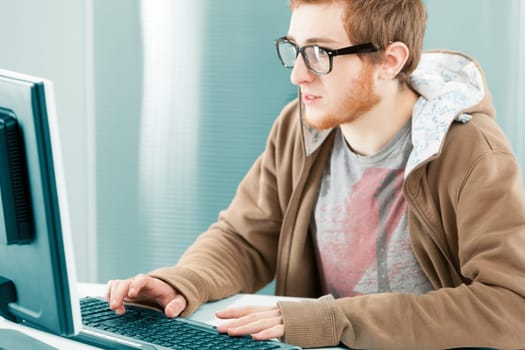 Young millennial worker in front of computer screen. Wearing black prescription glasses, a brown hoodie. He is typing and is extremely interested and focused.