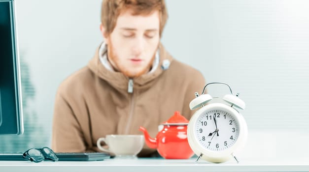 Close-up close-up in focus of white alarm clock on the table of a young worker out of focus in the background who is falling asleep in the office while eating breakfast. On the table a teapot and a te