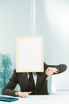 Sign-face man framed in an office. One hand is resting on the desk but the other makes a distinct thumbs-down gesture. Concept of negative judgment, rejection, wrong.