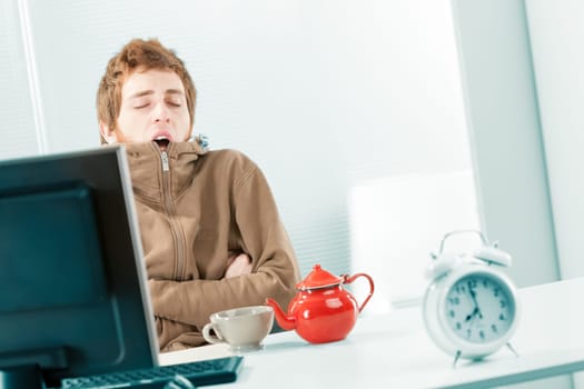 Dutch shot of a cold and sleepy, shivering young worker yawning as he huddles in his brown sweatshirt. He eats breakfast in the office with a mug and a teapot on the table near the computer. In the fo