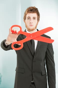 A black pinstriped office worker with a beard and short red hair holds a large red scissor. Cutting concept. Cutting staff, cutting costs and expenses. Hardly discounts: his expression is very serious