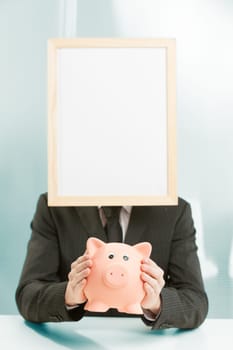 Office workplace, a sign-headed man framed in light wood holds a piggy bank. He holds it carefully, for a concept of savings, spending, investment and generally related to money and business economics