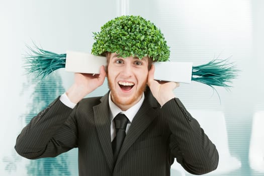 A clerk in an office makes a super smiling and elated face. He has the leafy foliage of a tree instead of hair and pretends to listen to two pots of grass like shells to hear the sound of the sea. Mad