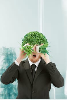 A clerk in an office makes a stunned face in a surreal mood. He has a bush for hair and looks through two pots of grass side by side as if through binoculars. 80s style in the mimicry. Black pinstripe