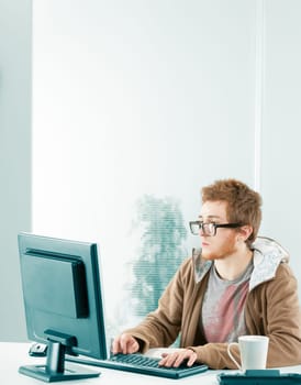 Wide shot with wide copysopace. Young nerdy millennial worker in front of computer screen. Wearing black prescription glasses, a brown hoodie. He is typing and is extremely interested and focused.