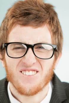Close-up frontal portrait of young man with short red hair and beard. Funny expression, grimacing, he makes beaver teeth by squinting his eyes and the reflection of light on the black frames of his gl