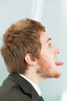 Close-up side portrait of young man with short red hair and beard in suit and tie. Facing to the right (but facing away from himself) he sticks out his entire tongue in defiance, mockery, and sneer at