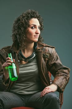Elegant chair portrait of a biker woman in leather jacket and goggles. She has a defiant look and holds a green glass bottle: enjoying her beer.