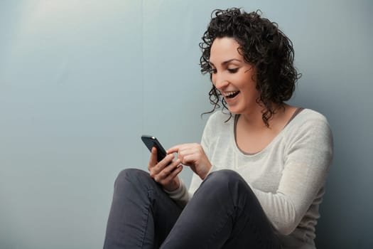 Woman on her home's floor, engrossed with her smartphone's digital content. From videos to social media, every entertainment form is accessible. Donning cozy winter wear like jeans and sweater, she be