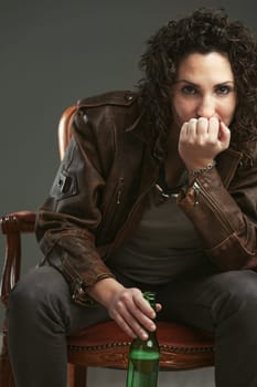 Elegant chair portrait of a biker woman in leather jacket and goggles. She has a defiant look and holds a green glass bottle: enjoying her beer.