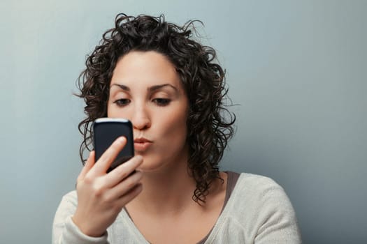 woman, with curly hair and a full figure, leans on her home wall. She blows a kiss towards her phone, possibly snapping a selfie or video chatting, clad in a sweater
